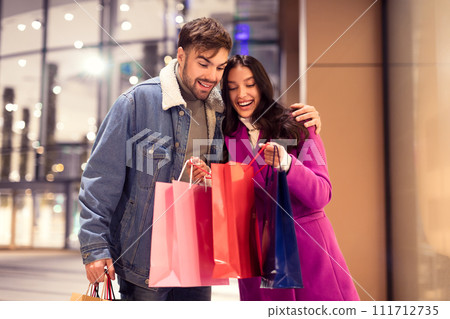 couple shopping peeking inside bags outside mall at winter night 111712735