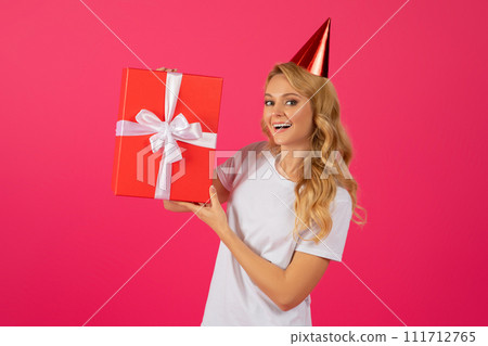 Happy blonde woman wearing birthday hat holding gift, pink background 111712765