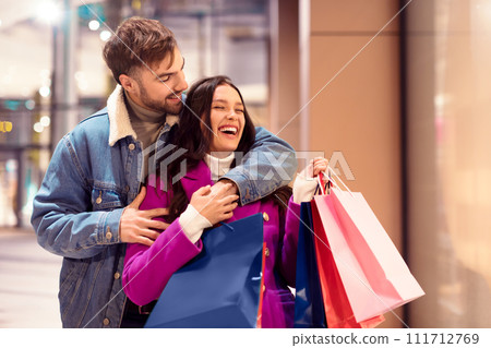 Cheerful buyers couple outside of store hugging with shopping bags 111712769
