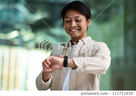 Smiling young asian man checks time on smartwatch outdoor Smiling young asian man checks time on smartwatch outdoor 111712839