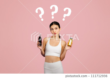 Slim european lady in sportswear holding bottle with smoothie and carbonated drink, thinking what to choose, pink background 111712936