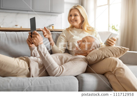 Happy senior couple sitting on sofa at home, using gadgets 111712958
