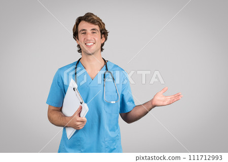 Excited male doctor in scrubs holding clipboard and showing something on empty palm pointing with hand at copy space 111712993