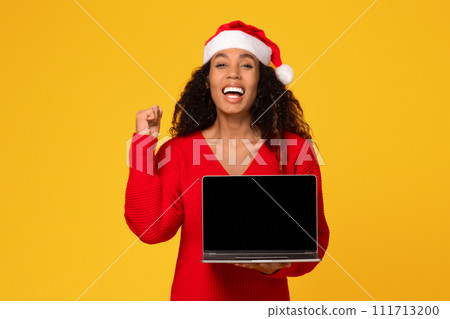 Overjoyed black woman in Santa hat showing laptop with blank screen and gesturing yes sign 111713200