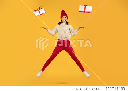 woman in knitted cap jumps with two presents, yellow background 111713463