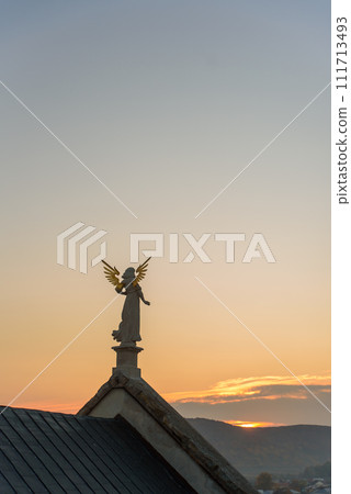 Stone sculpture of an angel with golden wings standing on the rooftop of a baroque temple at sunset. Stone sculpture of an angel with golden wings standing on the rooftop of a baroque temple at sunset. 111713493