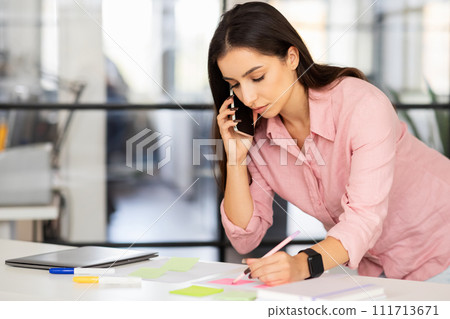 Busy focused young arab woman manager make notes at desk, call by phone Busy focused young arab woman manager make notes at desk, call by phone 111713671