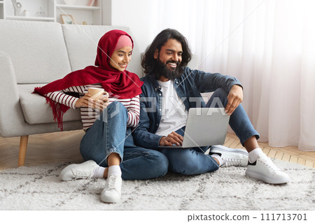 Relaxed young muslim couple sitting on floor with laptop and coffee mug 111713701