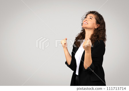 Elated young european businesswoman with beaming smile looks up, raising her fists in victorious gesture, free space 111713714