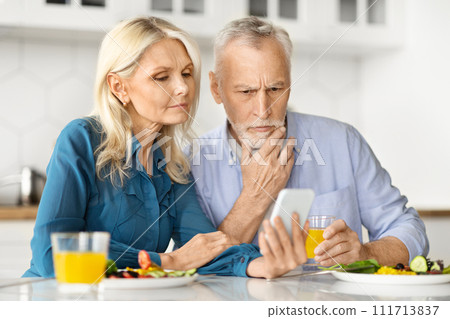 Worried Older Spouses Looking At Smartphone And Having Breakfast In Kitchen Worried Older Spouses Looking At Smartphone And Having Breakfast In Kitchen 111713837