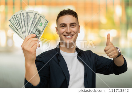 Smiling handsome millennial caucasian guy in casual, show many dollars cash, thumb up gesture 111713842