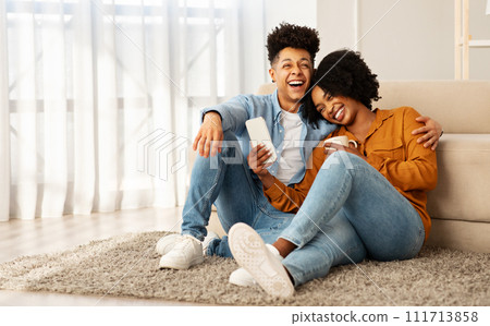 A joyful couple sitting on the floor against a couch, sharing a light-hearted moment with a smartphone 111713858