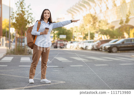 Cheerful pretty young caucasian woman tourist with backpack, cup of coffee takeaway, stop taxi 111713980