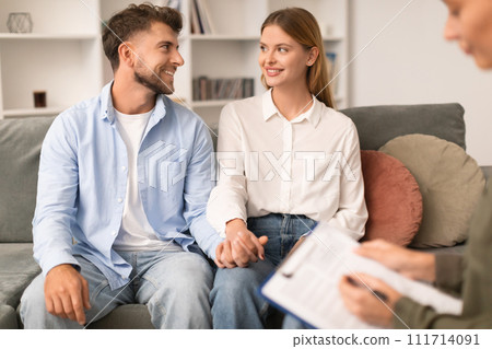 Cheerful millennial couple seated on therapist's couch sharing smiles 111714091