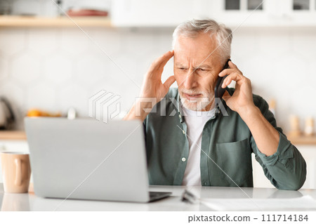 Stressed senior man using phone and laptop, sitting at desk in kitchen 111714184