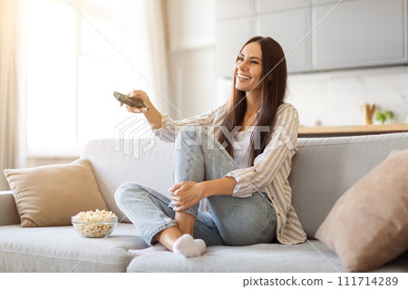 Smiling young woman watching TV and eating popcorn at home 111714289