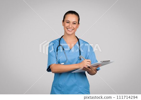 Smiling caucasian woman doctor surgeon, nurse in blue uniform, making notes at clipboard and smiling on gray background Smiling caucasian woman doctor surgeon, nurse in blue uniform, making notes at clipboard and smiling on gray background 111714294
