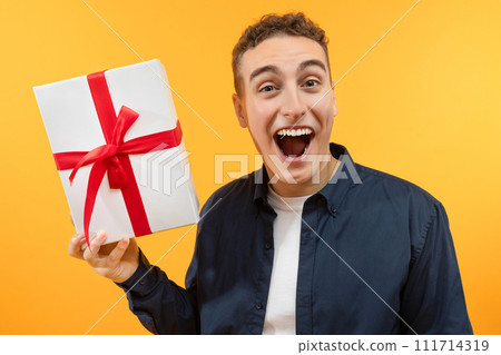 Happy young guy holding wrapped box, feeling excited on yellow Happy young guy holding wrapped box, feeling excited on yellow 111714319