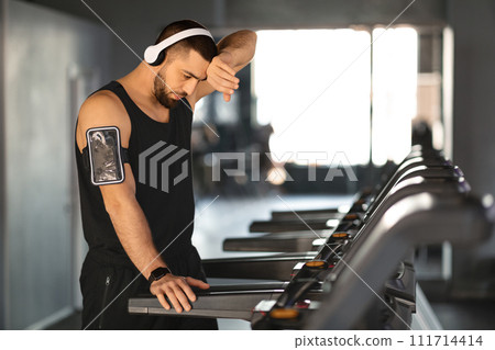 Exhausted young athlete wipes sweat from his forehead after treadmill session 111714414