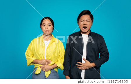 Concerned young woman with hands clasped while a man beside her winces with a hand on his stomach Concerned young woman with hands clasped while a man beside her winces with a hand on his stomach 111714505