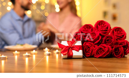 Gift box and roses on table with couple enjoying a romantic dinner 111714573