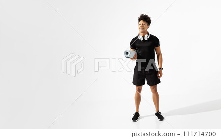 Athletic latin man with headphones holding fitness mat, white background 111714700