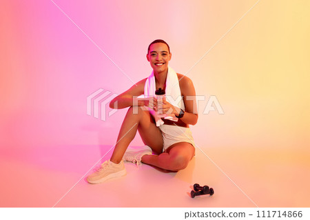 Happy young latin lady in sportswear sit with towel, bottle of water, resting from workout 111714866