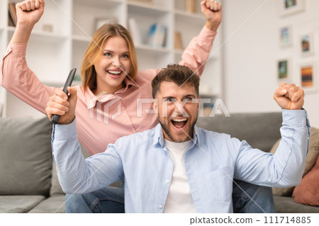 Excited Couple Watching TV And Shaking Fists In Joy Indoors 111714885