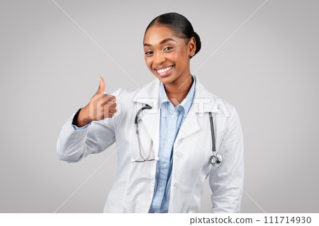 Cheerful pretty millennial black lady doctor in white coat make thumb up sign with hand Cheerful pretty millennial black lady doctor in white coat make thumb up sign with hand 111714930