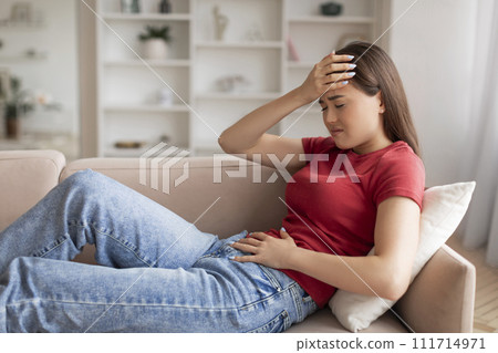 Worried asian woman suffering from stomach pain, holding her abdomen and head 111714971