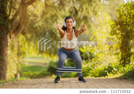 Young athletic lady doing squats with resistance band outdoors 111715032