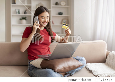 Smiling asian woman holding credit card and talking on cellphone at home Smiling asian woman holding credit card and talking on cellphone at home 111715347