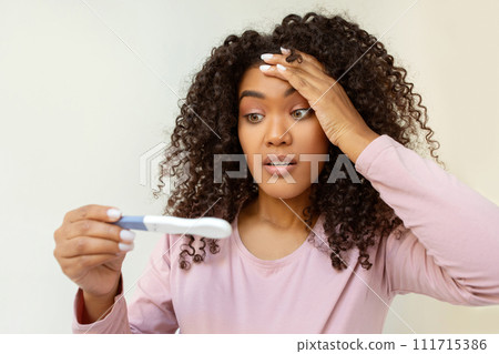 Surprised young black woman holding pregnancy test and looking with disbelief Surprised young black woman holding pregnancy test and looking with disbelief 111715386