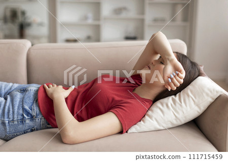 Young asian woman experiencing discomfort in stomach while lying on couch 111715459