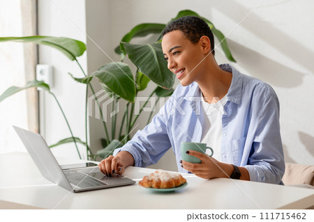 Smiling young latin lady enjoy cup of coffee and croissant at table and video call on laptop 111715462