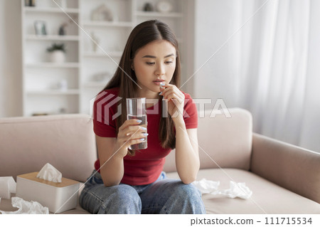 Concerned young asian woman taking pill with glass of water 111715534