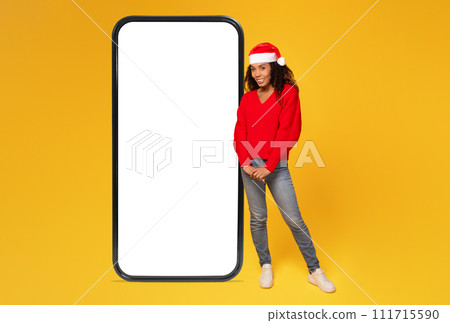Cheerful black lady in Santa hat standing near big phone with white blank screen 111715590