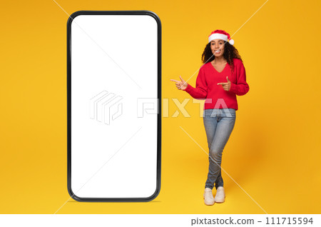 African american lady in Santa hat points at phone blank screen, full length, mockup African american lady in Santa hat points at phone blank screen, full length, mockup 111715594