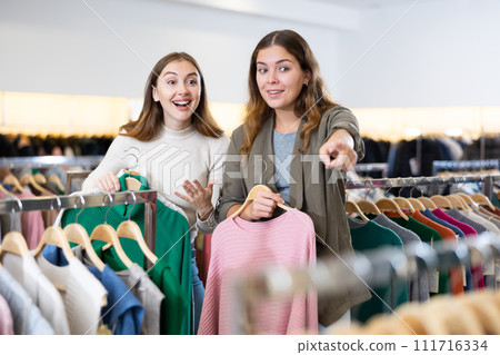 Amazed female holding warm clothes on hangers and showing her friend a discount during shopping in a store Amazed female holding warm clothes on hangers and showing her friend a discount during shopping in a store 111716334