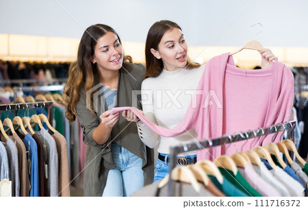 Cheerful female friends talking and chossing pink cardigan in a clothing store Cheerful female friends talking and chossing pink cardigan in a clothing store 111716372