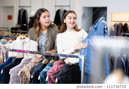Two cheerful female friends comparing warm white and blue gilets in a shopping mall Two cheerful female friends comparing warm white and blue gilets in a shopping mall 111716719