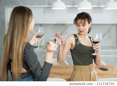 Two joyful middle-aged women dancing together at the kitchen-table with wineglasses 111716730