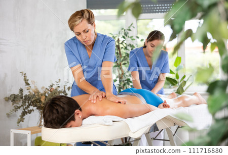 Two female masseur doing massage at back man who lying on massage table in beauty salon 111716880