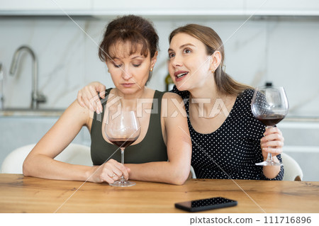 Middle-aged woman sitting at table while another woman trying to calm her sitting by her side Middle-aged woman sitting at table while another woman trying to calm her sitting by her side 111716896