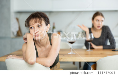 Upset middle-aged woman sitting at the kitchen table with her back to another lady quarreling to her 111717001