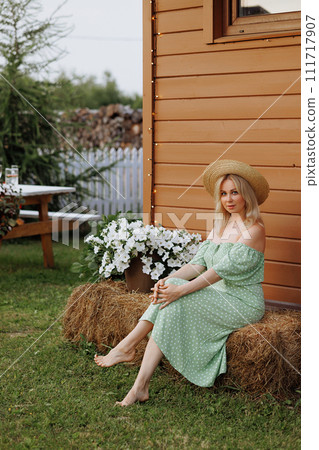 A woman in a green dress sits on hay bales near a wooden house A woman in a green dress sits on hay bales near a wooden house 111717907
