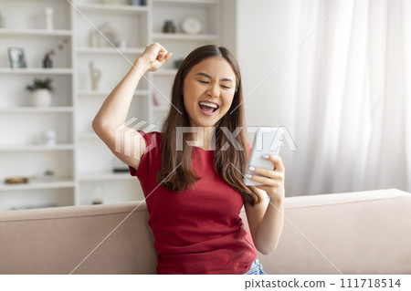 Euphoric young asian woman cheering success with smartphone at home 111718514