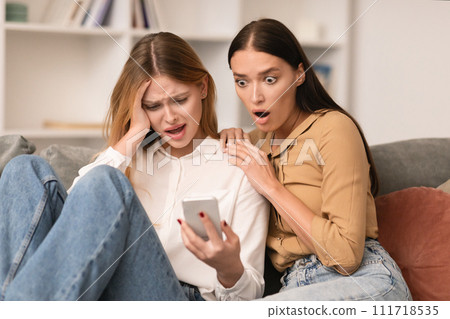woman shares shocking text message on smartphone with friend indoors woman shares shocking text message on smartphone with friend indoors 111718535