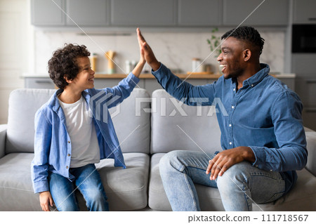 Joyful black father and son high-fiving, celebrating happy moment on the couch 111718567