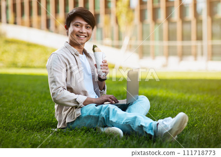 happy asian guy eating sandwich, working on laptop outdoors 111718773